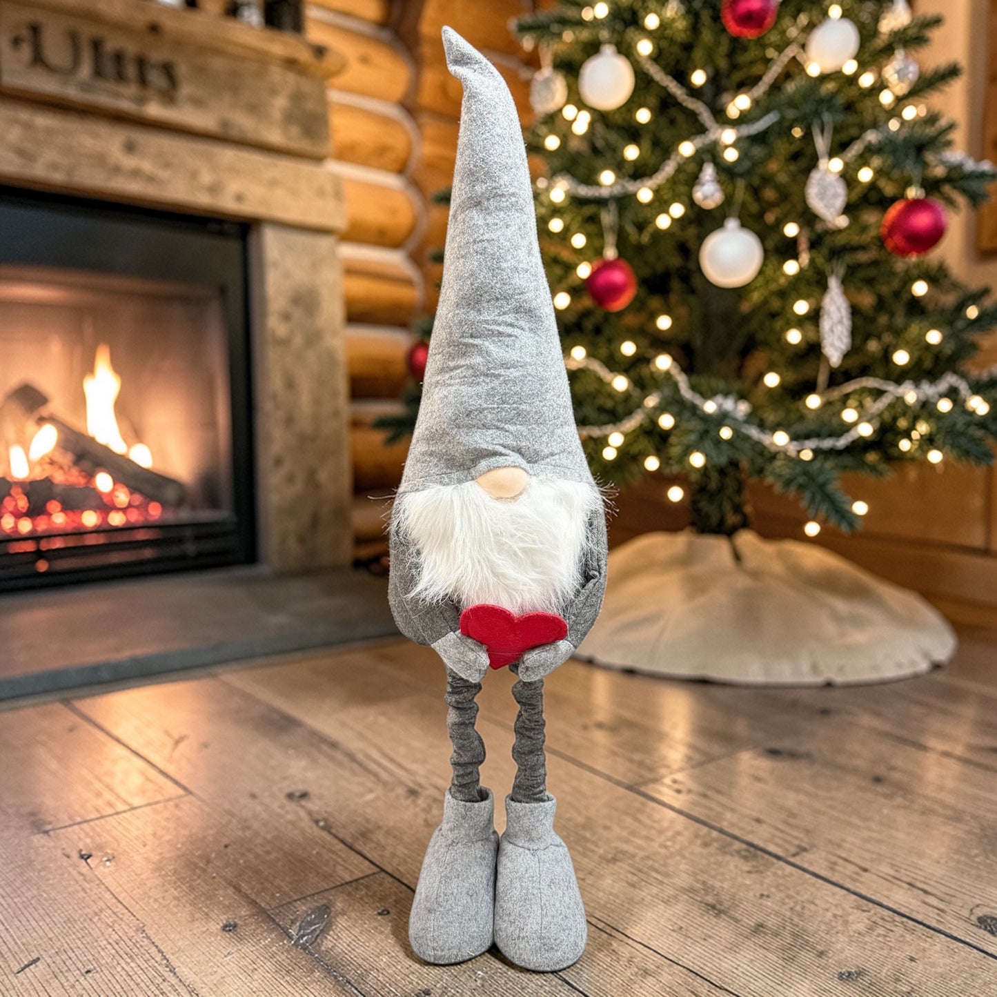 Graue Wichtelfigur aus Stoff mit langem Bart und rotem Herz in den Händen auf einem Holzboden vor einem Weihnachtsbaum und Kamin.