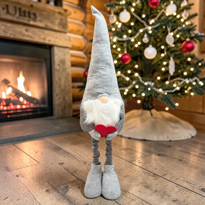 Graue Wichtelfigur aus Stoff mit langem Bart und rotem Herz in den Händen auf einem Holzboden vor einem Weihnachtsbaum und Kamin.