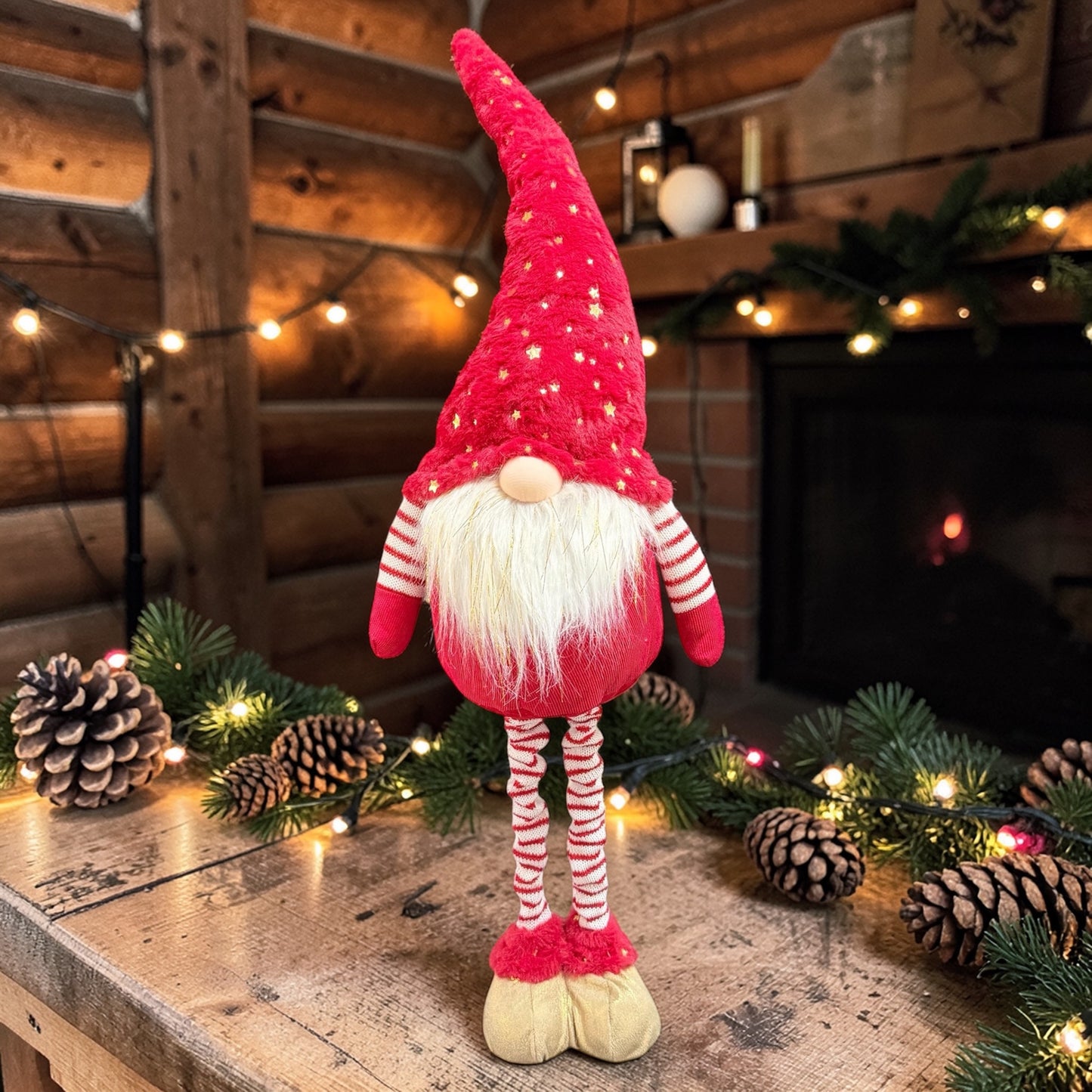 Rote Wichtelfigur aus Stoff mit weißem Bart auf einer Holzfläche mit Tannenzweigen, Lichterkette und Kamin im Hintergrund.