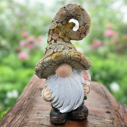 Wichtel mit weißem Bart, naturfarbener Mütze und rosa Blume in der Hand, liebevolle Gartendekoration im Grünen.