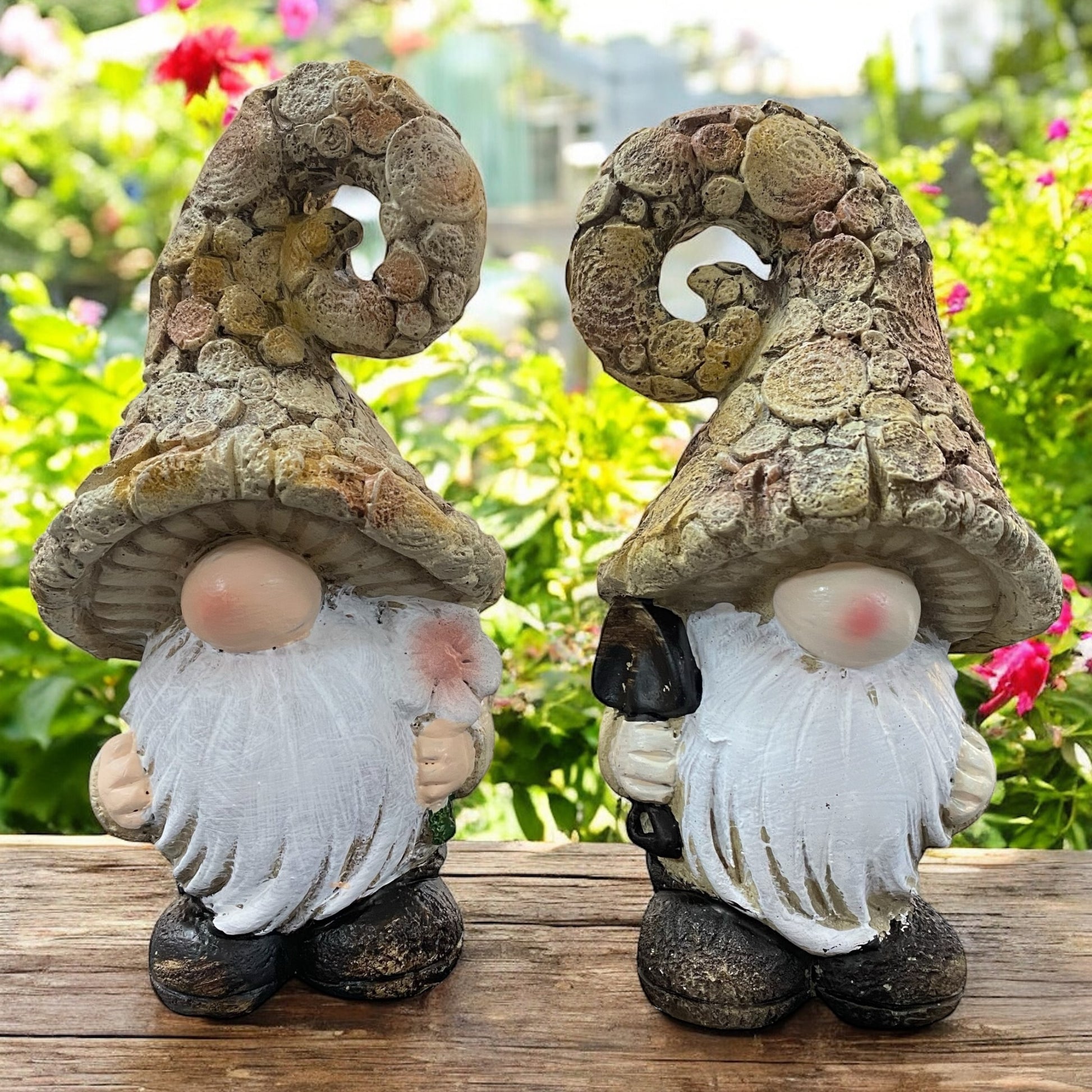 Zwei naturfarbene Wichtel-Figuren mit Bart und gedrehten Mützen, dekorativ im Garten zwischen Blumen arrangiert.