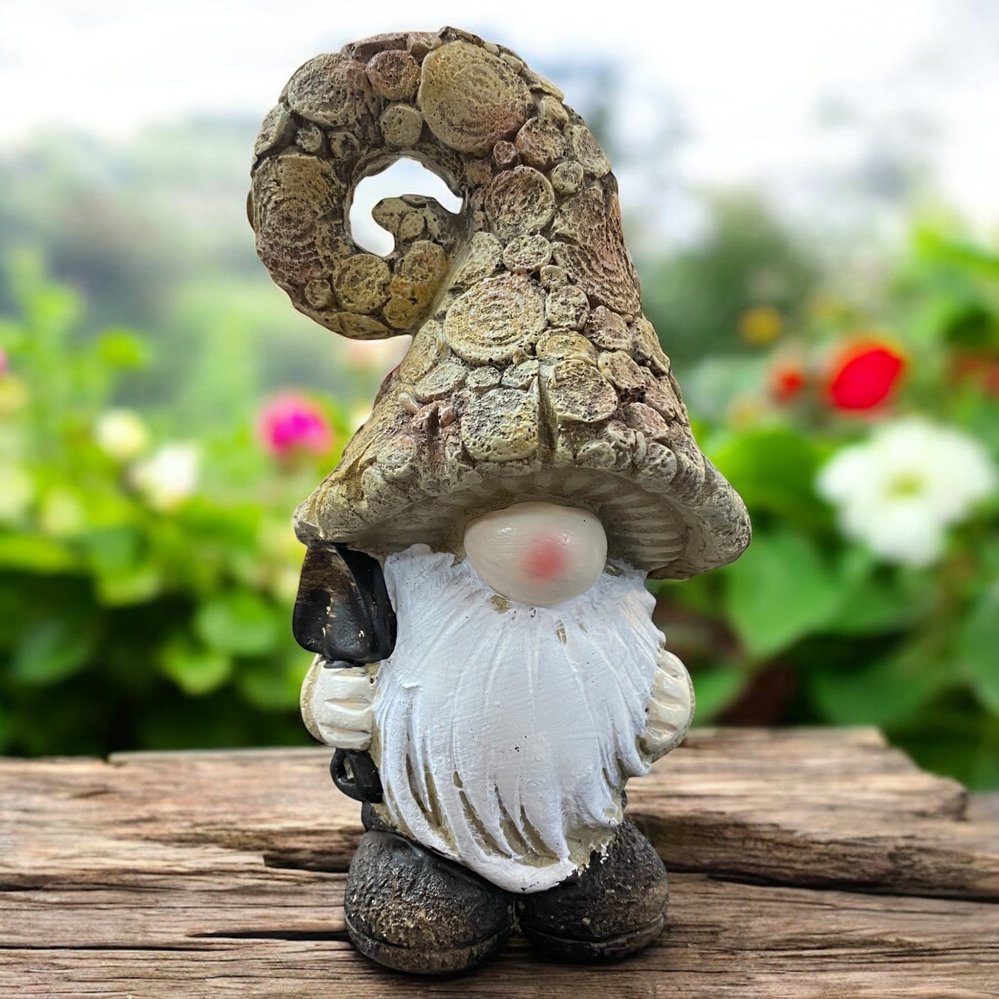 Wichtel mit naturfarbener, gedrehter Mütze und Schaufel in der Hand, rustikale Gartendeko auf Holztisch mit Blumenhintergrund.
