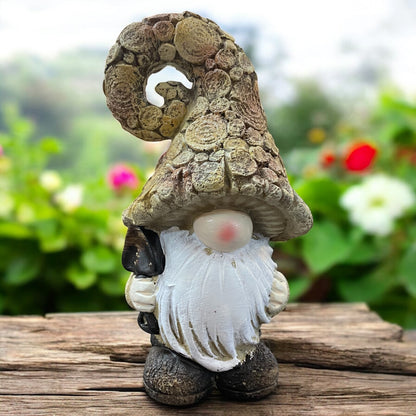 Wichtel mit naturfarbener, gedrehter Mütze und Schaufel in der Hand, rustikale Gartendeko auf Holztisch mit Blumenhintergrund.
