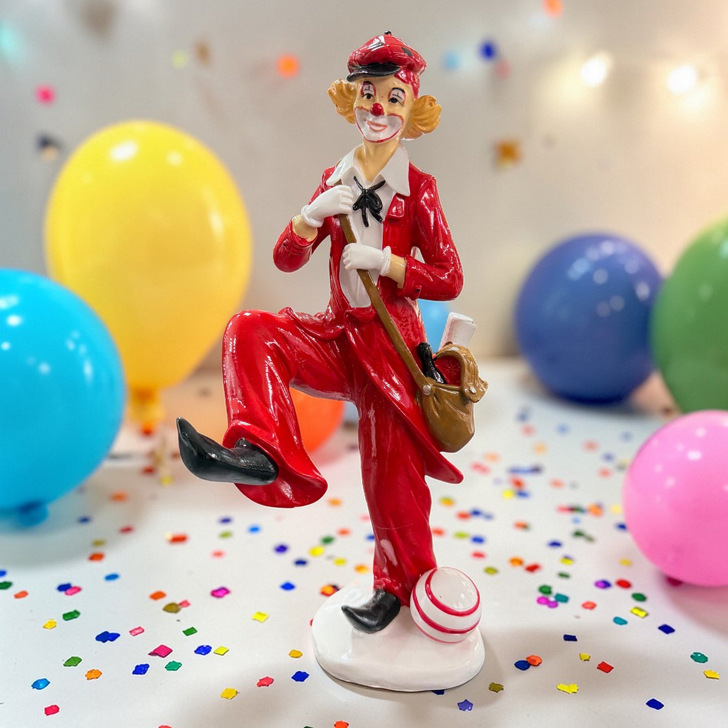 Clown-Dekofigur in Rot mit Umhängetasche und Ball vor Luftballons und Konfetti.
