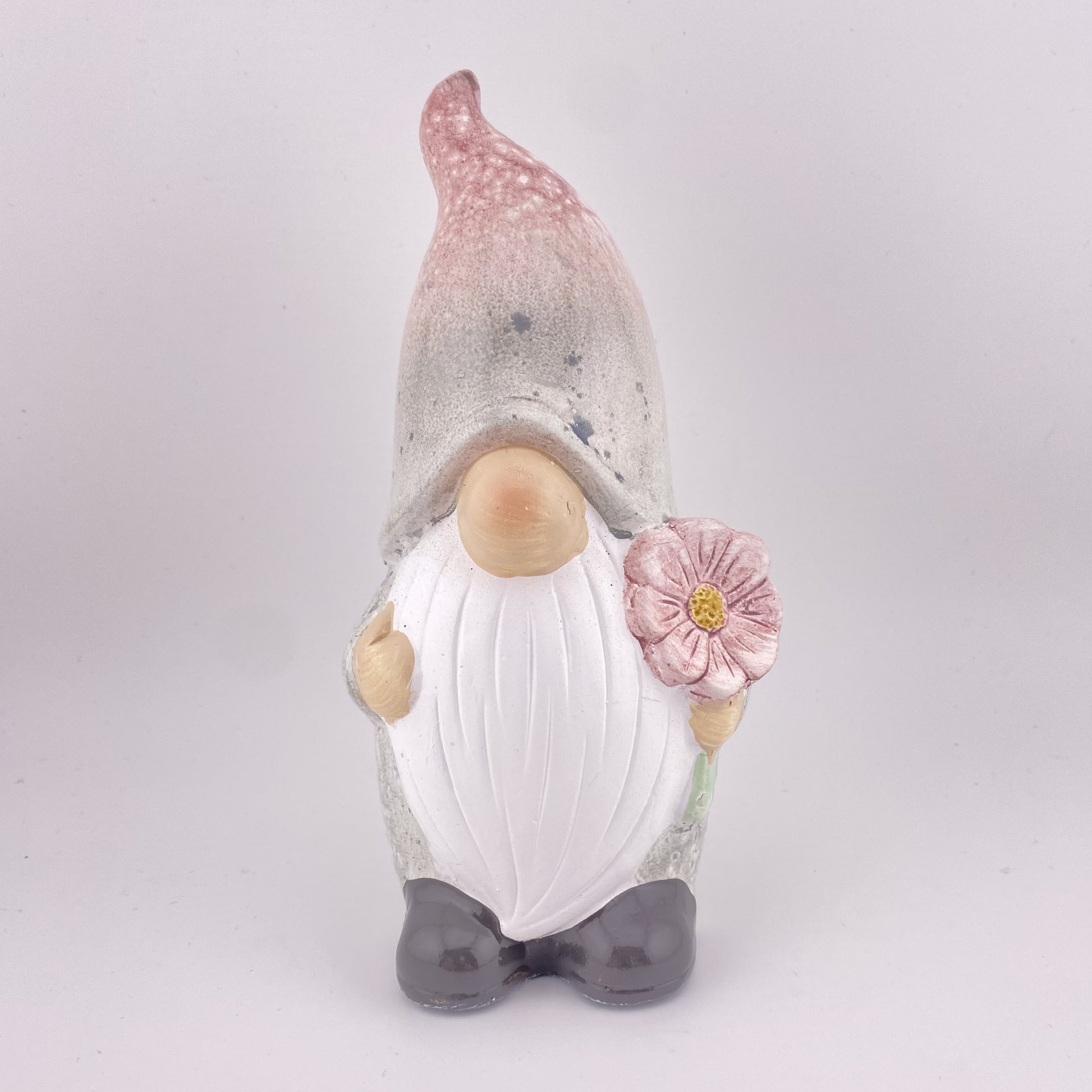 Ein Deko Wichtel Fabelwesen mit Zipfelmütze, weißem Bart und Blume in der Hand ist vor weißem Hintergrund platziert. 