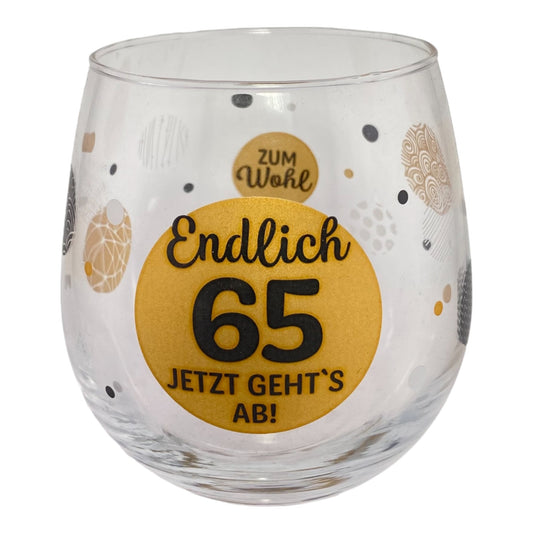 Glas mit Schriftzug "Endlich 65 Jetzt gehts ab. Zum Wohl" vor weißem Hintergrund