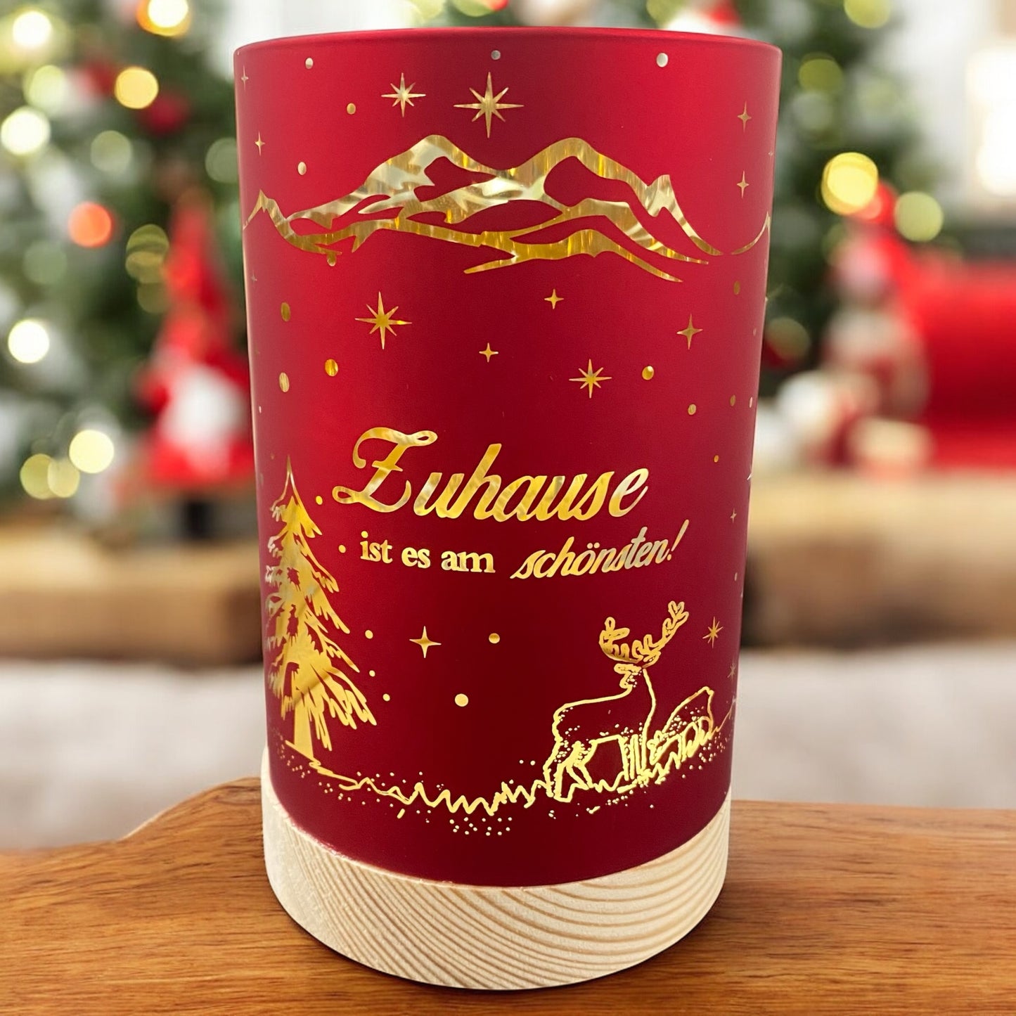 Kleines rotes Deko-Licht mit goldener Aufschrift „Zuhause ist es am schönsten!“ und leuchtender Weihnachtslandschaft vor festlich geschmücktem Hintergrund.