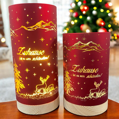 Zwei rote Deko-Lichter mit goldener Winterlandschaft und dem Schriftzug „Zuhause ist es am schönsten!“, auf hellem Holzsockel im Ambiente