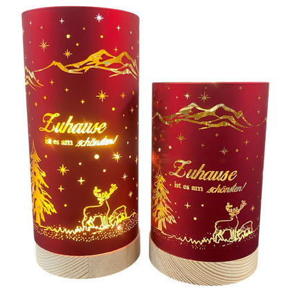 Zwei rote Deko-Lichter mit goldener Winterlandschaft und dem Schriftzug „Zuhause ist es am schönsten!“, auf hellem Holzsockel
