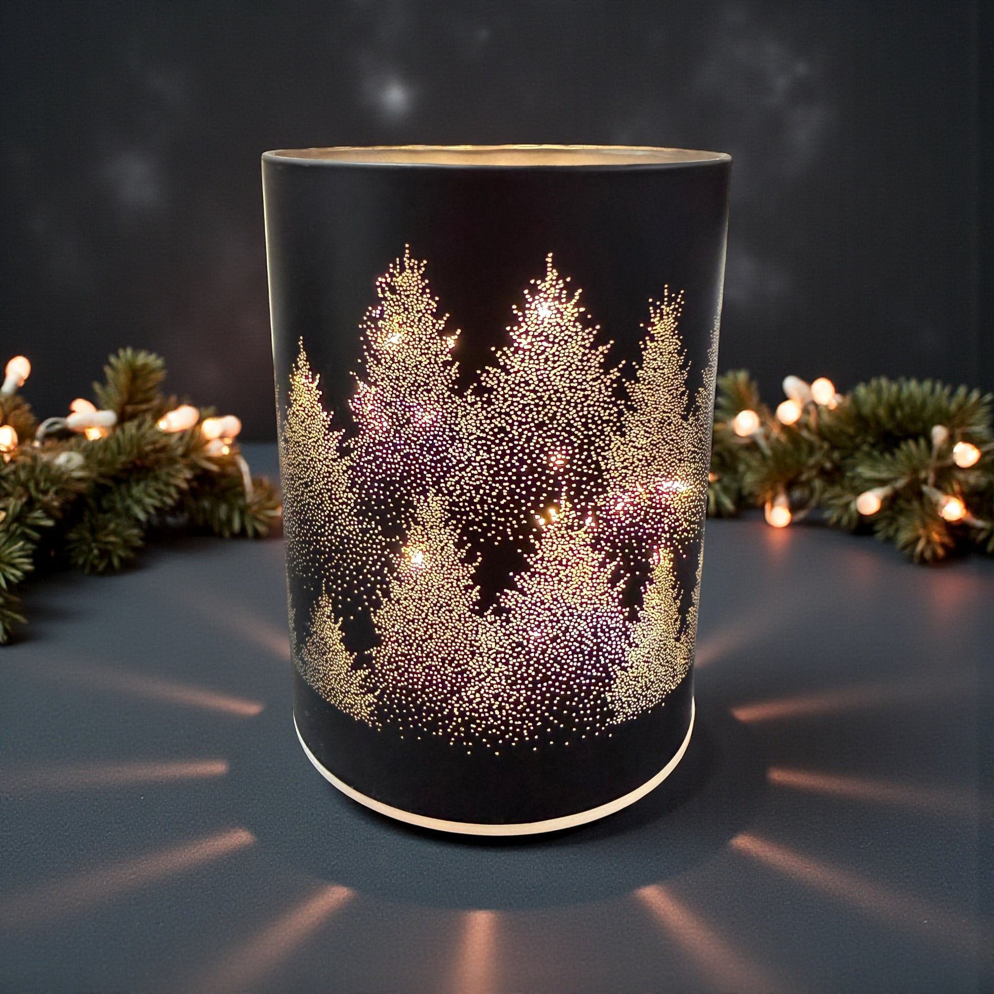 Ein zylindrisches schwarzes Dekolicht mit perforiertem Tannenbaum-Motiv und hellem Innenlicht steht auf einer Oberfläche vor Tannenzweigen und einer Lichterkette.