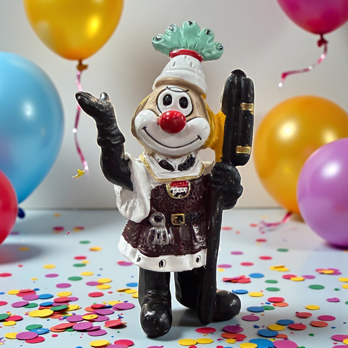 Clown-Figur in dunklem Kostüm mit Kopfbedeckung, eine Hand erhoben und ein längliches Objekt haltend, auf einer Tischfläche mit Konfetti und Luftballons im Hintergrund