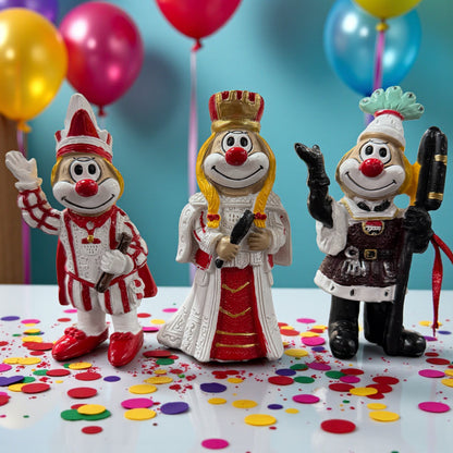 Dreigestirn bunte Clown-Figuren in unterschiedlichen Kostümen stehen nebeneinander auf einer Tischfläche mit Konfetti, im Hintergrund farbige Luftballons