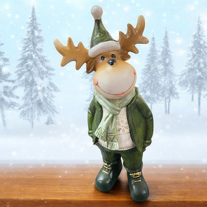 Weihnachtliche Elchfigur in grüner Kleidung mit Mütze und Schal, stehend auf Holzuntergrund vor winterlicher Schneelandschaft.