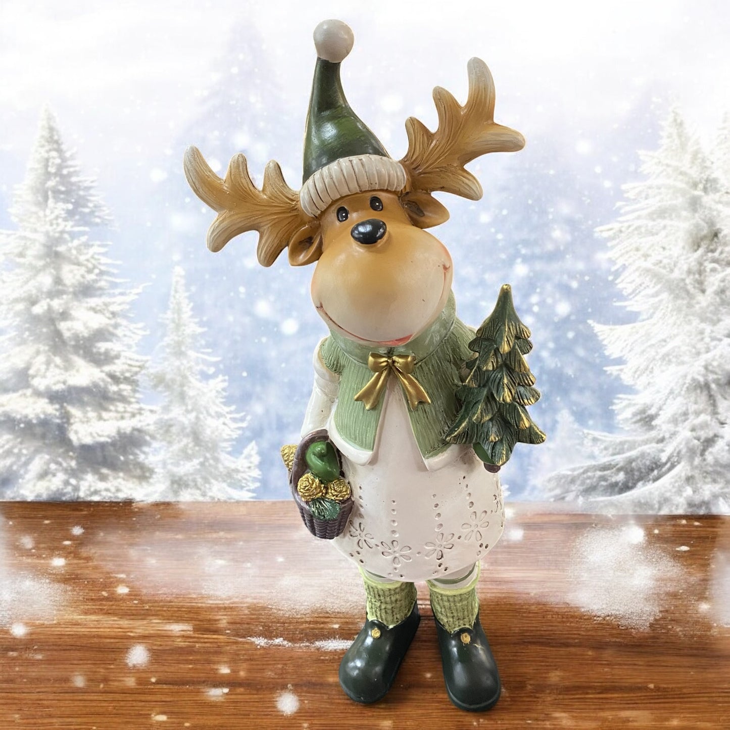 Große Elchfigur mit Geweih, grüner Mütze und Schal, hält ein Vogelhaus und steht auf Holzuntergrund vor verschneitem Waldhintergrund.