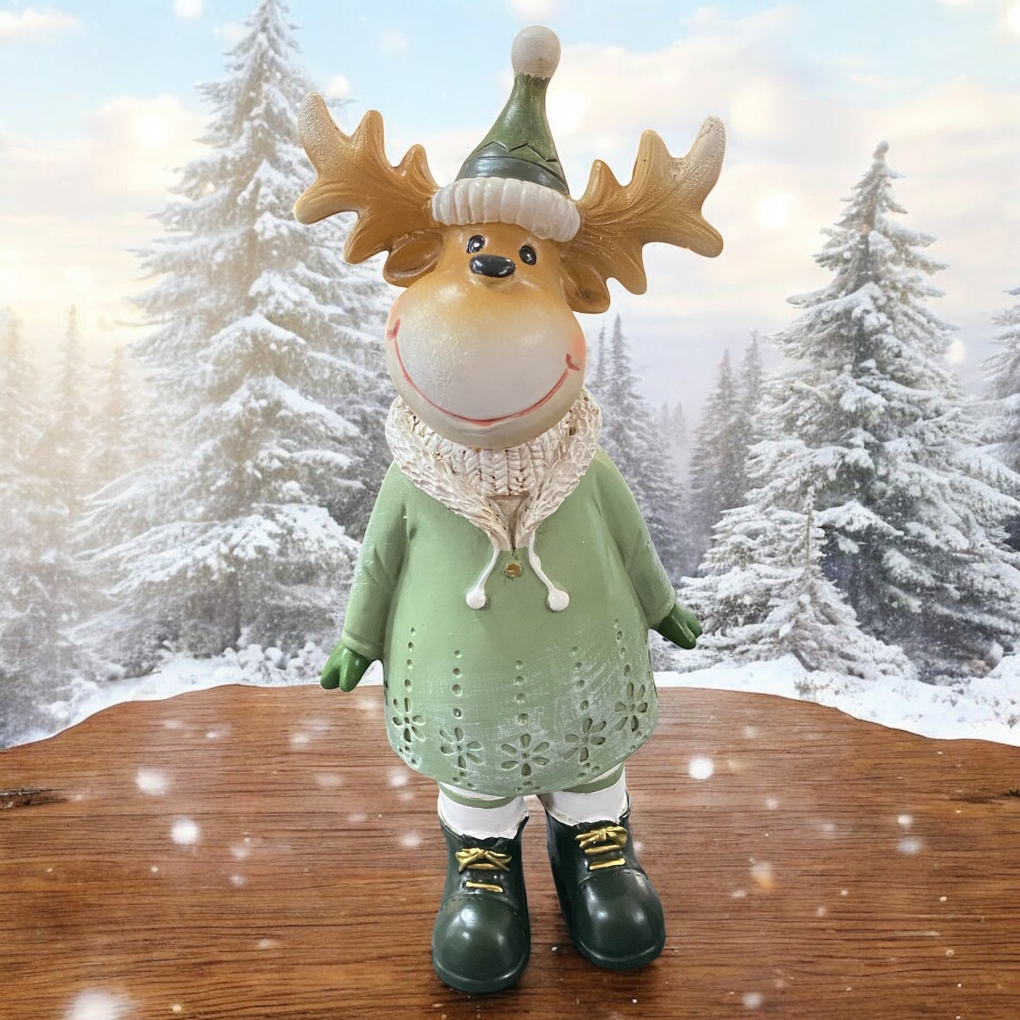 Kleine Elchfigur mit Geweih und grüner Mütze, trägt ein grünes Kleid und steht auf einem Holztisch vor verschneiter Winterlandschaft.