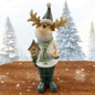 Weihnachtliche Elchfigur mit Geweih und grüner Mütze, trägt Kleid, Schal und hält einen kleinen Tannenbaum vor winterlicher Schneelandschaft.