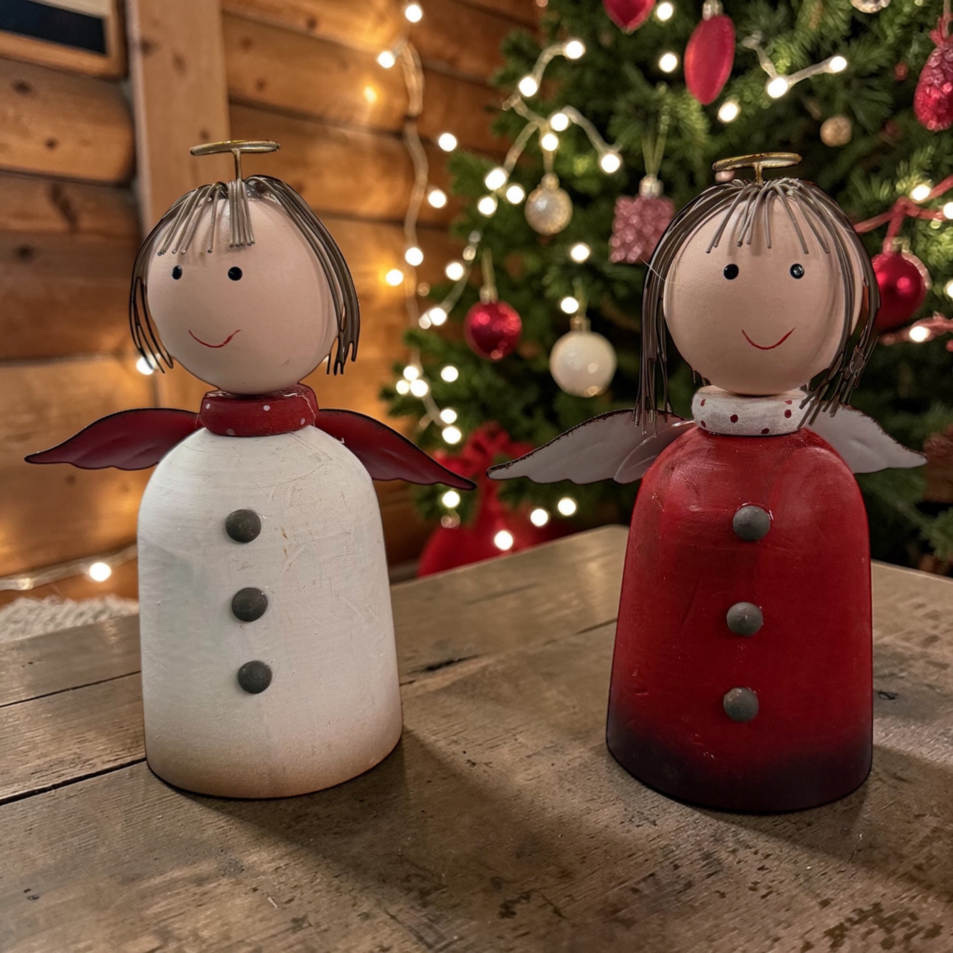 Zwei Engel-Dekofiguren in Rot und Weiß mit grauen Flügeln, auf einer Holzoberfläche vor Hintergrund mit Weihnachtsbaum, grünen Tannenzweigen und warmen gelblichen Lichtern.