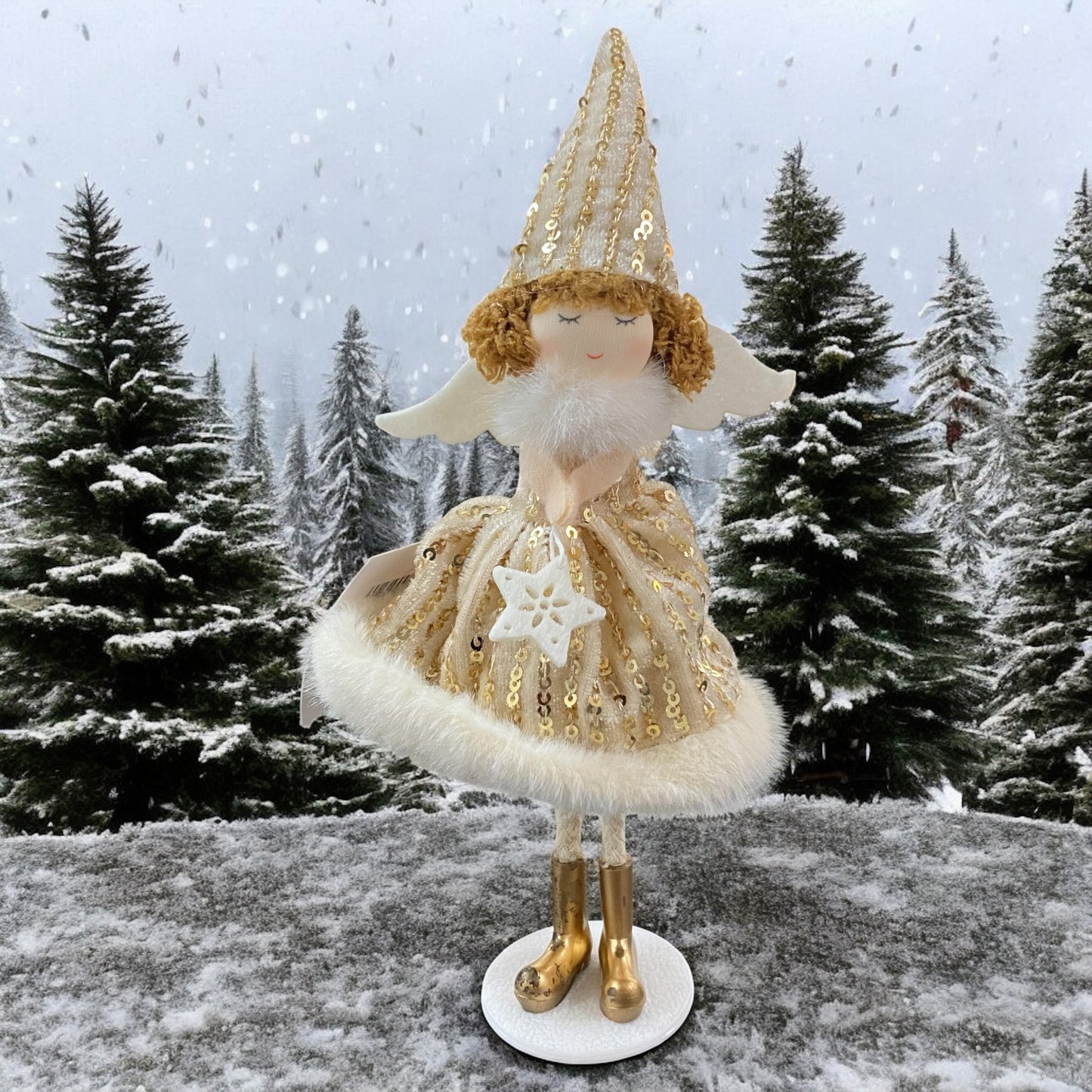 Stehende Stoff-Engelfigur mit beigem Paillettenkleid, weißen Flügeln und Mütze auf einem weißen Sockel, im Hintergrund verschneite Bäume.