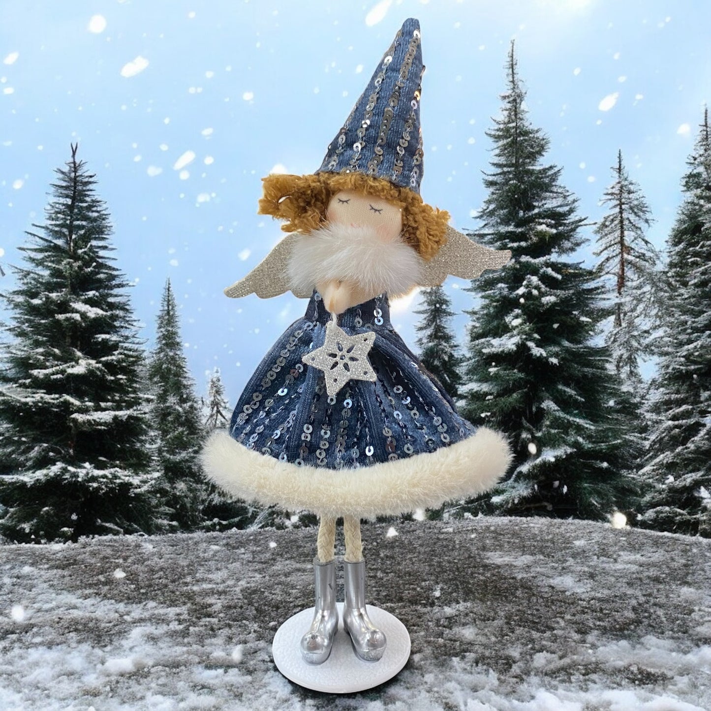 Stehende Stoff-Engelfigur mit blauem Paillettenkleid, silbernen Flügeln und Mütze auf einem weißen Sockel, im Hintergrund verschneite Bäume.