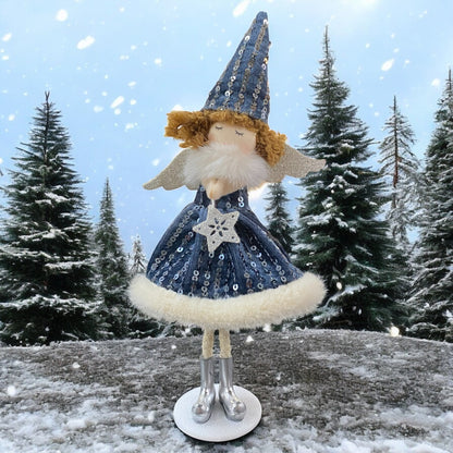Stehende Stoff-Engelfigur mit blauem Paillettenkleid, silbernen Flügeln und Mütze auf einem weißen Sockel, im Hintergrund verschneite Bäume.