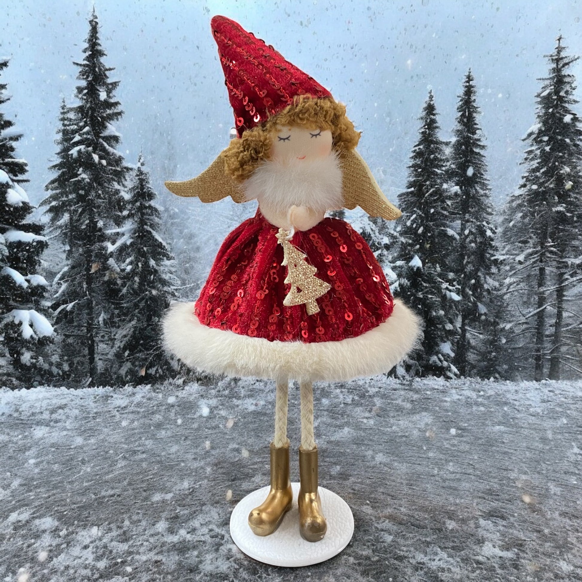 Stehende Stoff-Engelfigur mit rotem Paillettenkleid, goldenen Flügeln und Mütze auf einem weißen Sockel, im Hintergrund verschneite Bäume.

