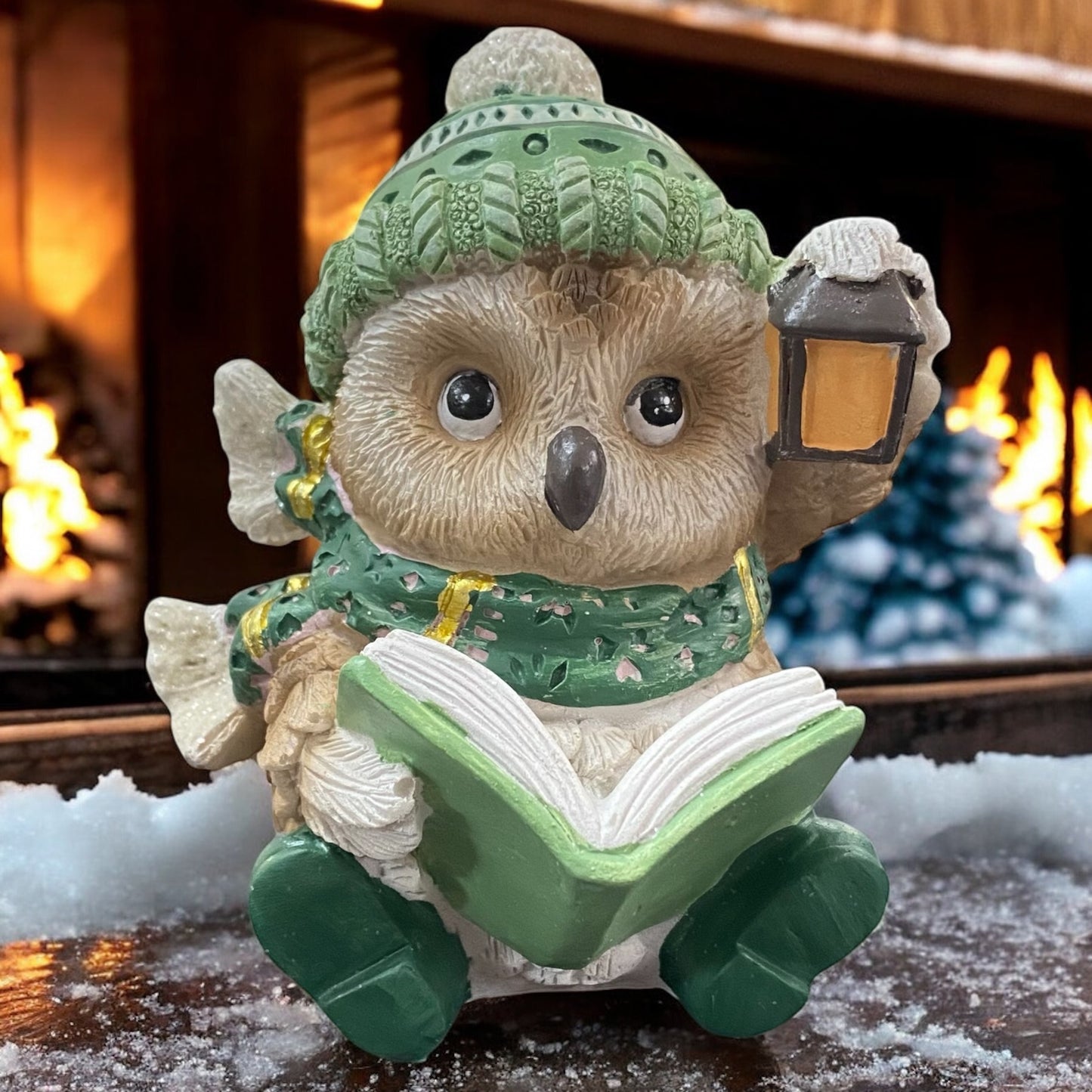 Winterliche Deko-Eule mit grüner Mütze, Schal und Buch in der Hand, vor Kaminfeuer arrangiert.