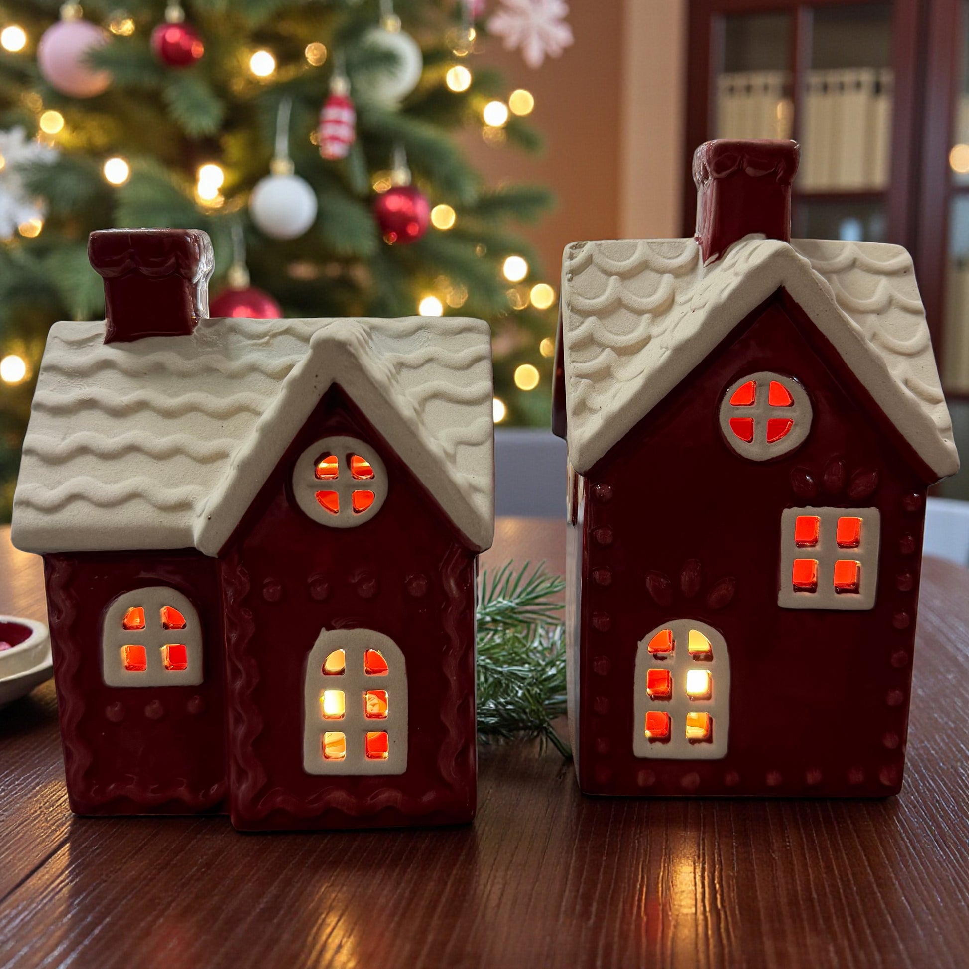 Dekohaus in roter Farbe mit weißem, verschneitem Dach und integrierter LED-Beleuchtung, durch deren Fenster warmes Licht scheint, platziert auf Holztisch vor Weihnachtsbaum mit Lichterkette und Kugeln im Hintergrund.