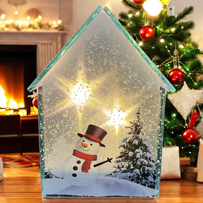 Beleuchtetes Glashaus mit Schneemann- und Tannenbaum-Motiv, steht auf einem Holztisch vor Kamin und Weihnachtsbaum.