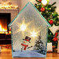 Beleuchtetes Glashaus mit Schneemann- und Tannenbaum-Motiv, steht auf einem Holztisch vor Kamin und Weihnachtsbaum.