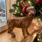 Hochlandrind-Figur mit roter Weihnachtsmütze. Position auf einer Holzfläche neben einem dekorierten Tannenbaum mit Kugeln und Lichtern. Hintergrund unscharf mit Lichteffekten.
