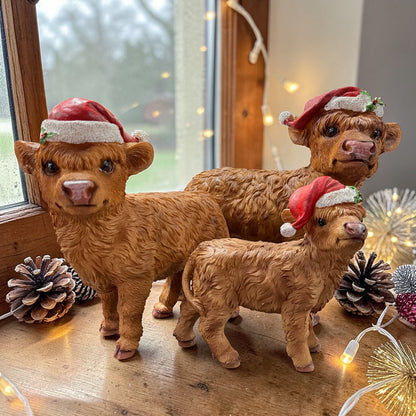 Drei Hochlandrind-Figuren mit Weihnachtsmützen auf einer Holzoberfläche nahe einem Fenster. Umgebung mit Lichterkette, Tannenzweigen und Zapfen. Warmes, dekoratives Ambiente.