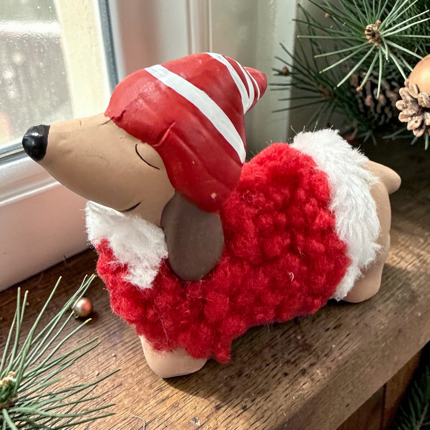 Einzelne Dackelfigur in liegender Position auf einer Holzoberfläche nahe einem Fenster. Rote Mütze und flauschiger roter Mantel mit weißen Details. Umgebung mit Tannenzweigen und weihnachtlicher Dekoration
