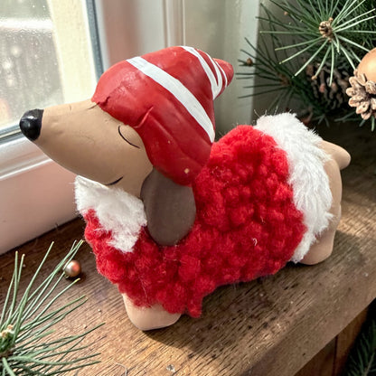 Einzelne Dackelfigur in liegender Position auf einer Holzoberfläche nahe einem Fenster. Rote Mütze und flauschiger roter Mantel mit weißen Details. Umgebung mit Tannenzweigen und weihnachtlicher Dekoration