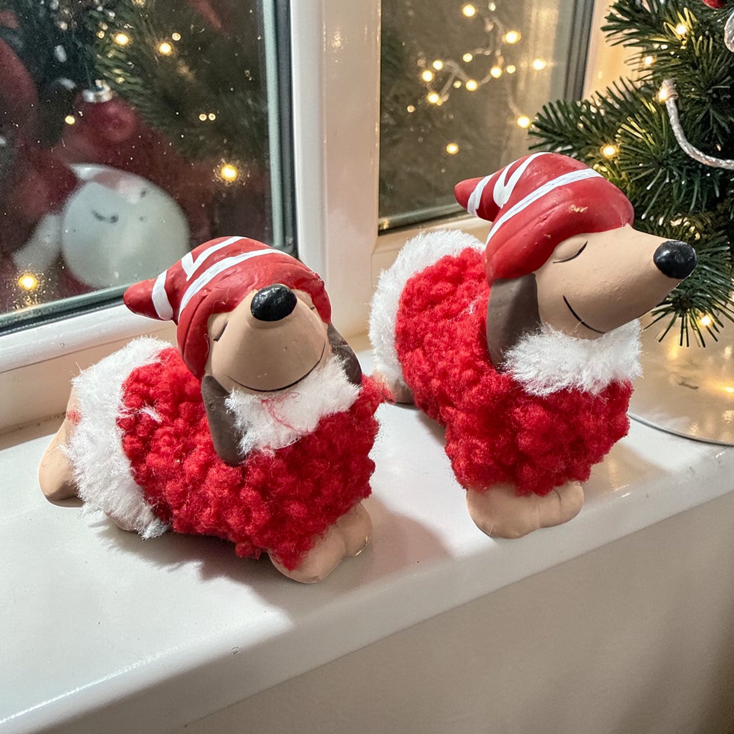 Zwei Dackelfiguren mit roter Mütze und flauschigem rotem Mantel auf einer Fensterbank. Im Hintergrund ein Fenster mit Lichterdekoration und weihnachtlichen Elementen wie Tannenzweigen.