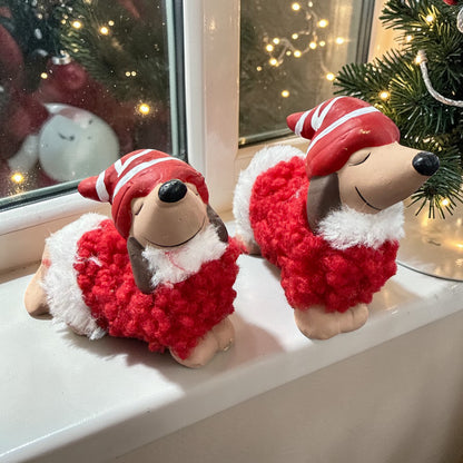 Zwei Dackelfiguren mit roter Mütze und flauschigem rotem Mantel auf einer Fensterbank. Im Hintergrund ein Fenster mit Lichterdekoration und weihnachtlichen Elementen wie Tannenzweigen.