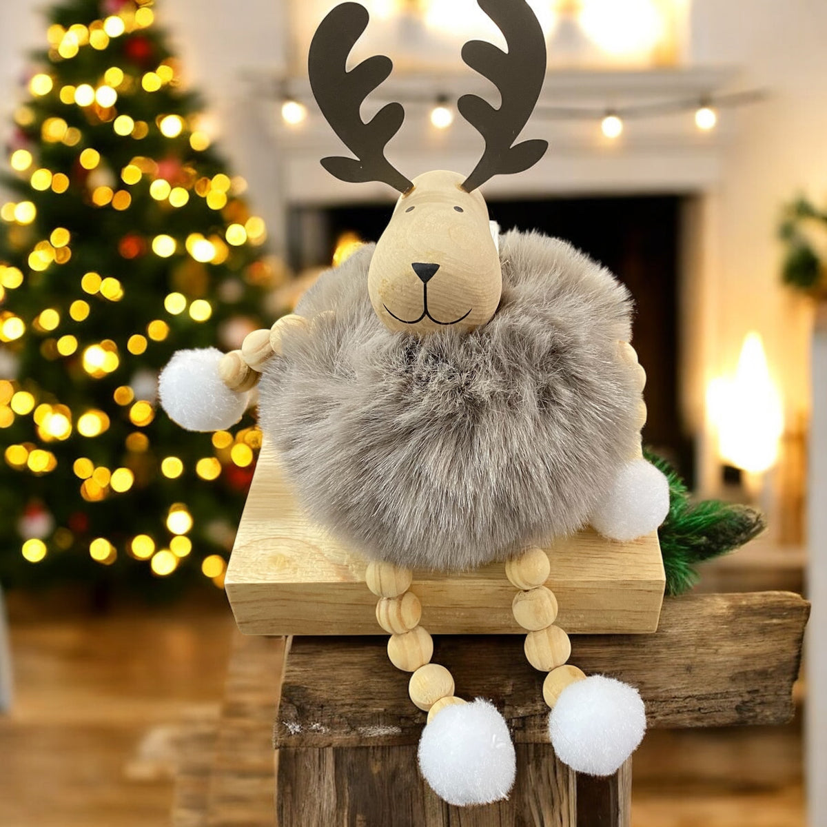 Sitzende Elchfigur mit braunen Geweihen und grauem, flauschigem Körper auf einem Holzblock vor weihnachtlich beleuchtetem Hintergrund.