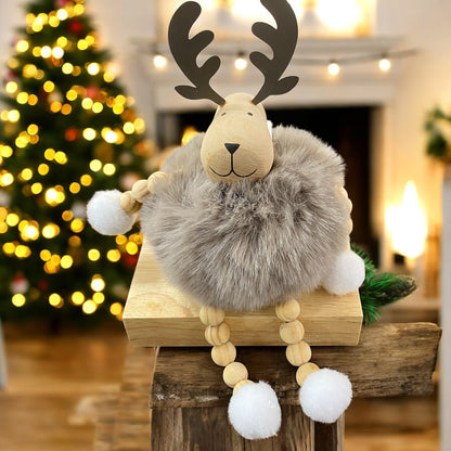 Sitzende Elchfigur mit braunen Geweihen und grauem, flauschigem Körper auf einem Holzblock vor weihnachtlich beleuchtetem Hintergrund.