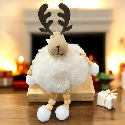 Sitzende Elchfigur mit braunen Geweihen und cremefarbenem, flauschigem Körper auf einem Holzblock vor Kamin- und Weihnachtsdekoration