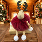 Sitzende Elchfigur mit braunen Geweihen und rotem, flauschigem Körper auf einem Holzblock vor weihnachtlich dekoriertem Hintergrund.