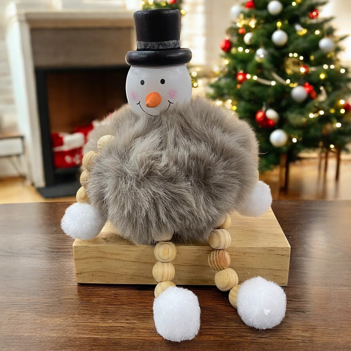 Sitzende Schneemannfigur mit schwarzem Hut und grauem, flauschigem Körper auf einem Holzblock vor Weihnachtsbaum und Kamin.