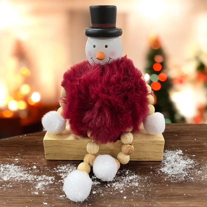 Sitzende Schneemannfigur mit schwarzem Hut und rotem, flauschigem Körper auf einem Holzblock vor weihnachtlicher Dekoration.