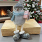 Winterliche Vogel-Figur mit Mütze und blauem Schal, die ein rosa Geschenk hält, auf einem Holzblock vor Kamin und geschmücktem Weihnachtsbaum.