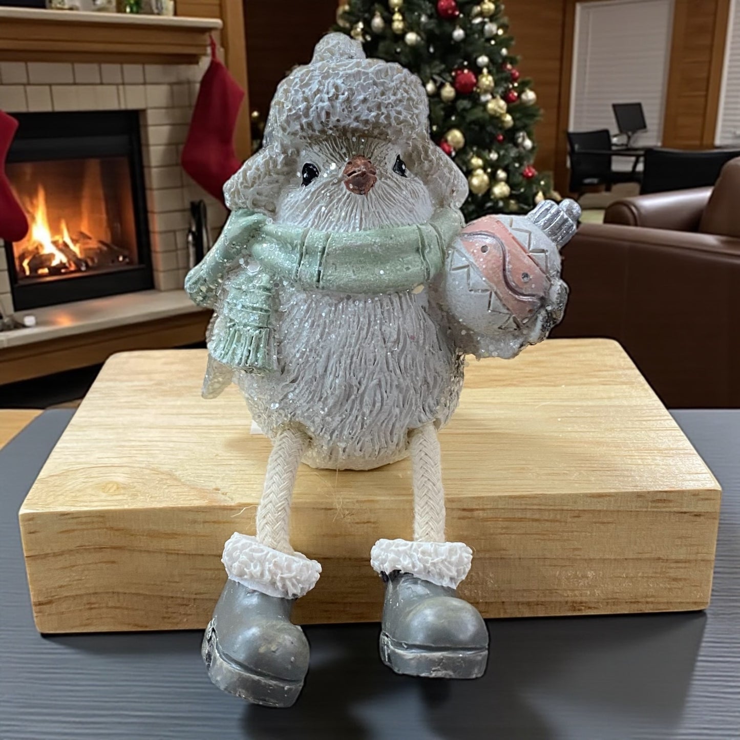 Winterliche Vogel-Figur mit Mütze und grünem Schal, die eine Weihnachtskugel hält, auf einem Holzblock vor Kamin und Weihnachtsbaum.