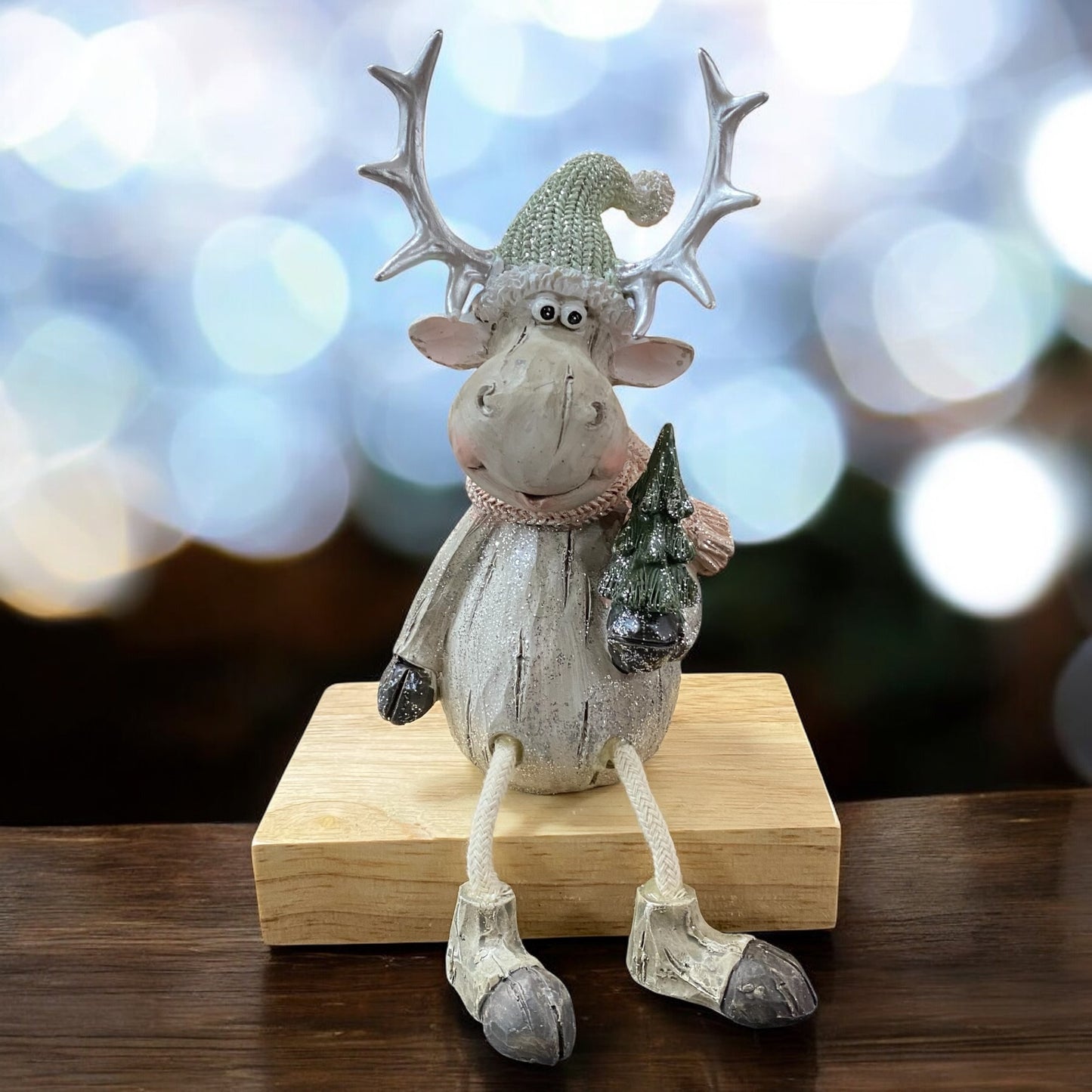 Sitzende Elchfigur mit grünem Strickmütze und Schal, hält einen kleinen Weihnachtsbaum, auf einem Holzblock vor festlichem Hintergrund.