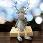 Sitzende Elchfigur mit grünem Strickmütze und Schal, hält einen kleinen Weihnachtsbaum, auf einem Holzblock vor festlichem Hintergrund.