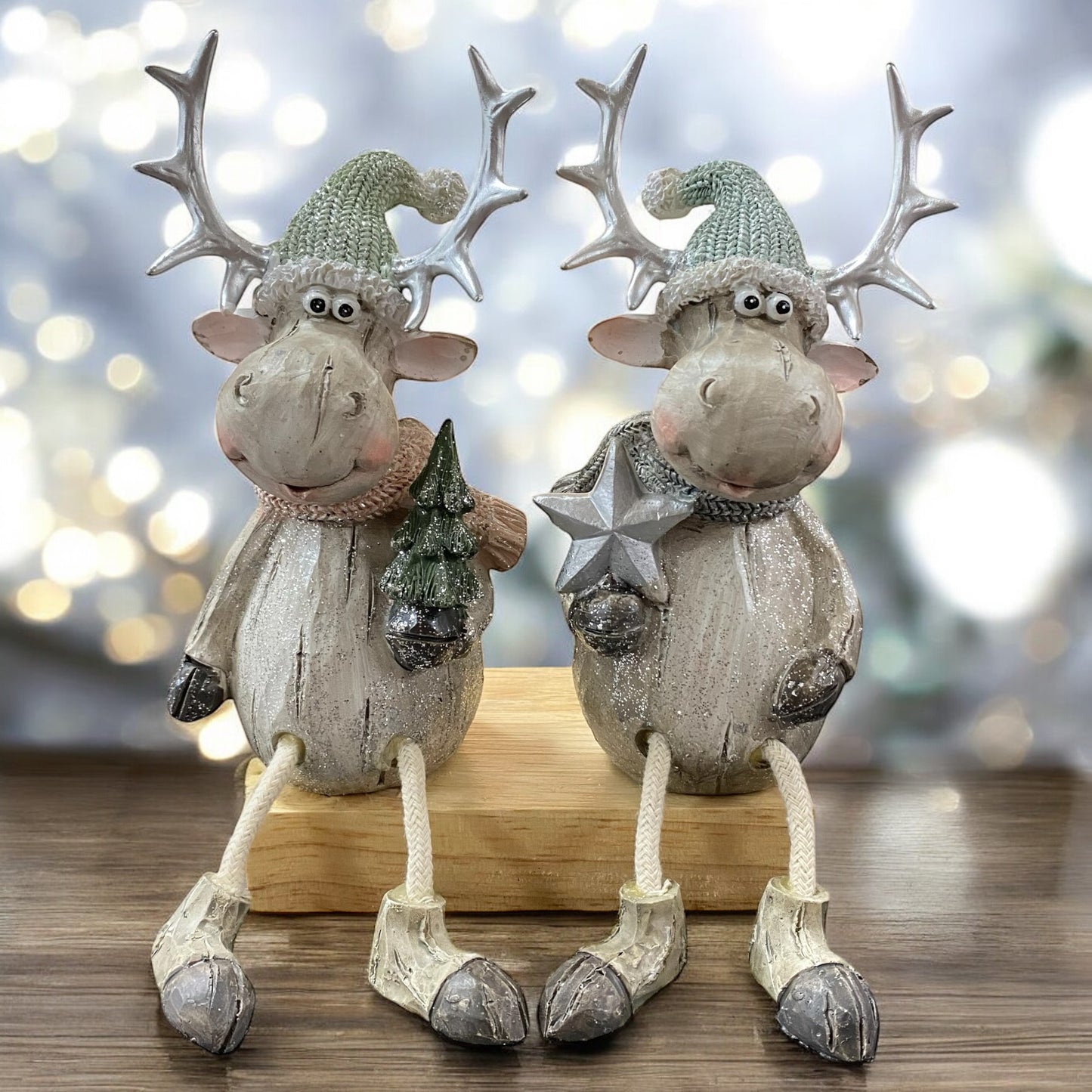 Zwei sitzende Elchfiguren mit großen Geweihen und grauen Mützen, eine hält einen Stern, die andere einen Tannenbaum, auf einem Holzblock im Ambiente