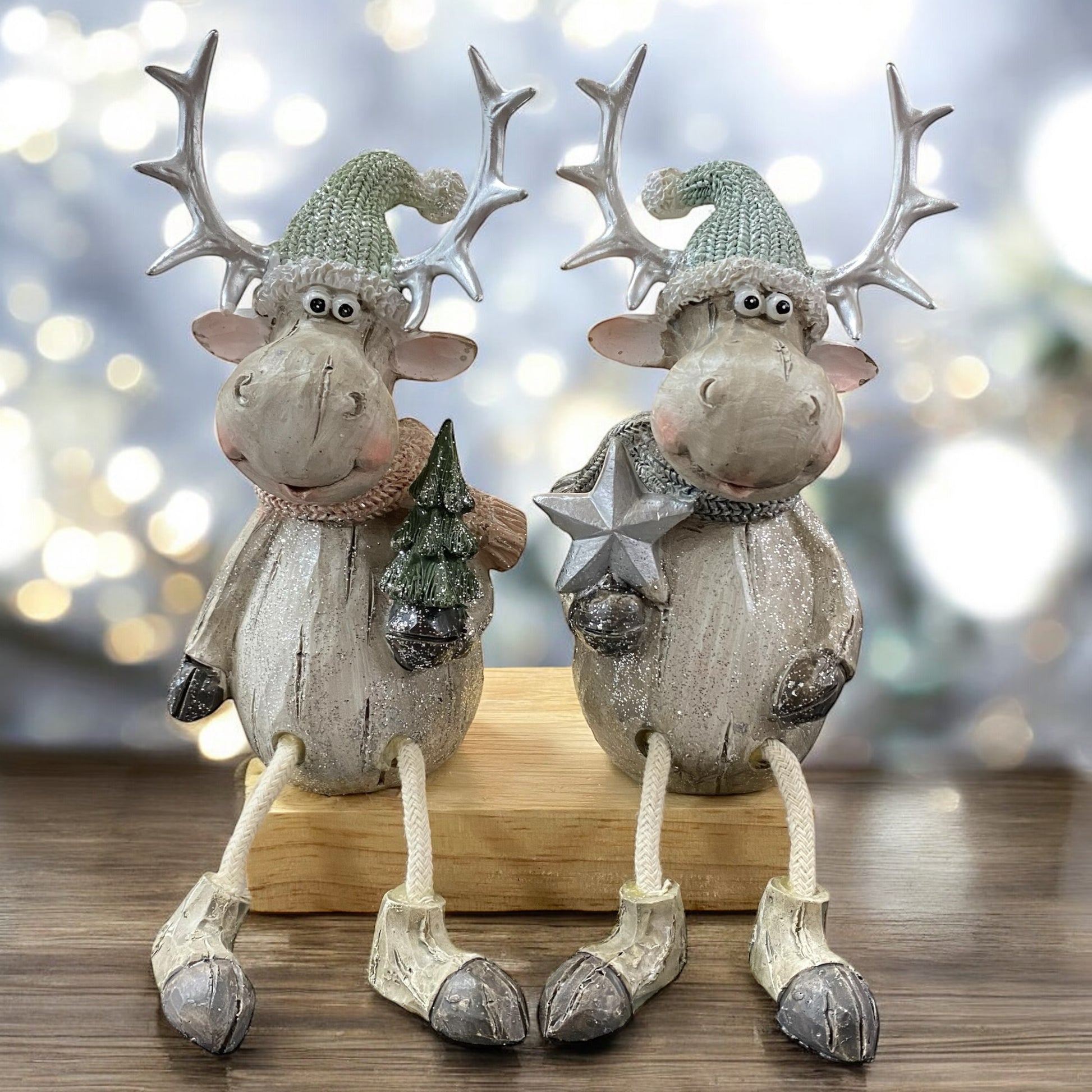Zwei sitzende Elchfiguren mit großen Geweihen und grauen Mützen, eine hält einen Stern, die andere einen Tannenbaum, auf einem Holzblock im Ambiente