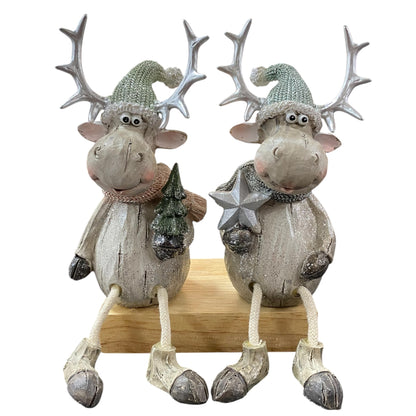 Zwei sitzende Elchfiguren mit großen Geweihen und grauen Mützen, eine hält einen Stern, die andere einen Tannenbaum, auf einem Holzblock.