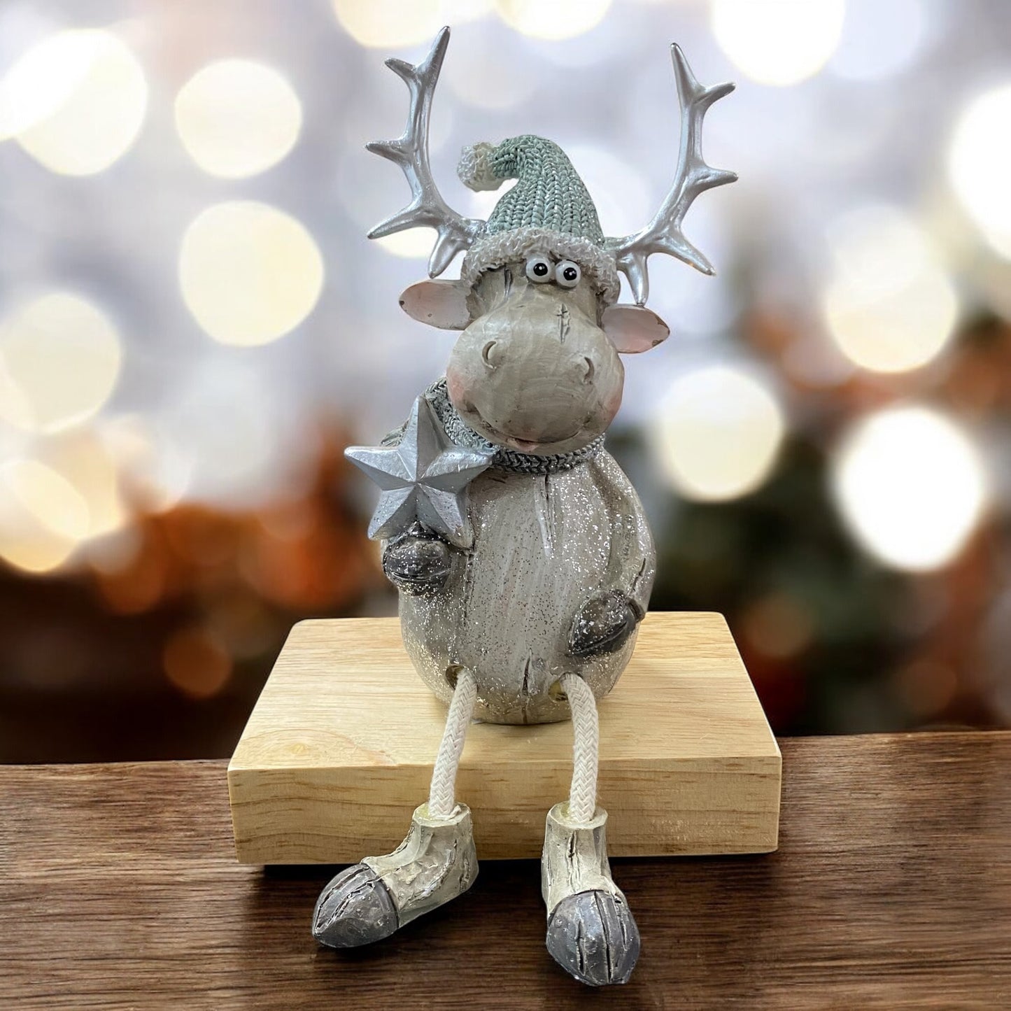 Sitzende Elchfigur mit grünem Strickmütze und Schal, hält einen silbernen Stern, auf einem Holzblock vor weihnachtlich beleuchtetem Hintergrund.
