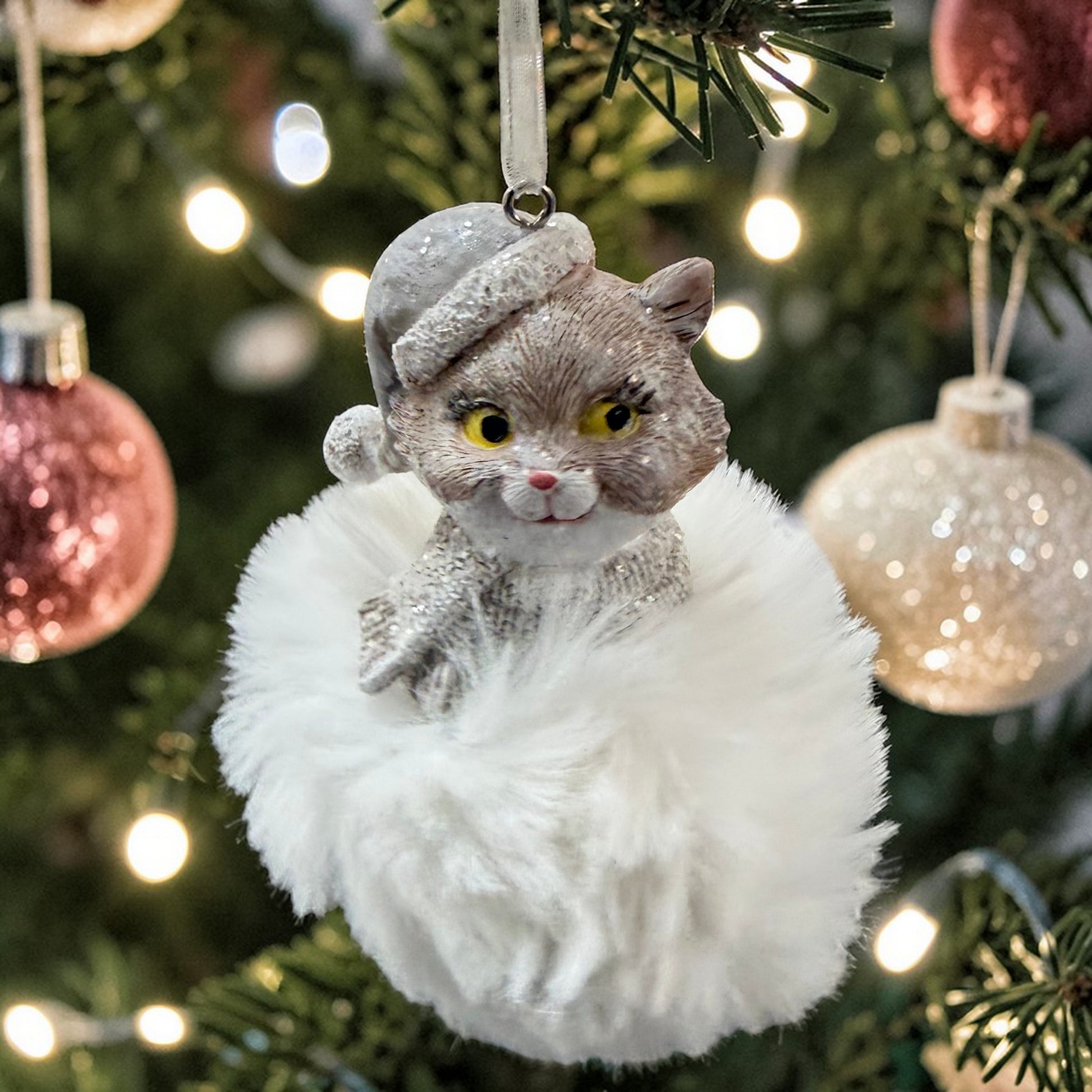 Ein Katzen-Anhänger mit Mütze auf einer weichen Plüschwolke hängt am festlich geschmückten Weihnachtsbaum.